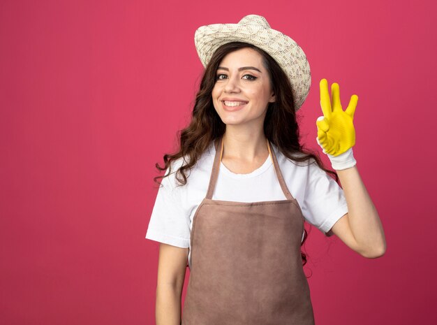 Sorridente giovane giardiniere femminile in uniforme che indossa guanti e cappello da giardinaggio gesti segno giusto della mano isolato sulla parete rosa con lo spazio della copia