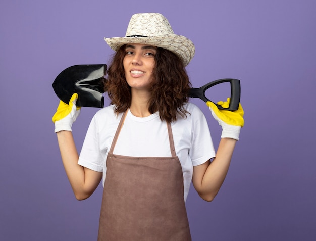 Sorridente giovane giardiniere femminile in uniforme che indossa guanti e cappello da giardinaggio tenendo la vanga sulle spalle