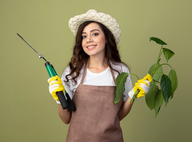 Sorridente giovane giardiniere femminile in uniforme che indossa guanti e cappello da giardinaggio tiene piante e tosatrici da giardino isolate sulla parete verde oliva con lo spazio della copia