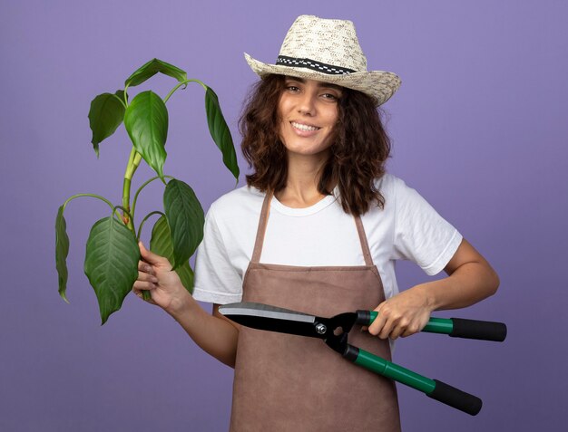 Sorridente giovane giardiniere femminile in uniforme che indossa il cappello da giardinaggio azienda pianta con clippers