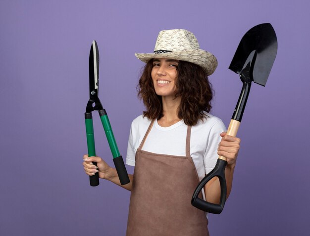 Sorridente giovane giardiniere femminile in uniforme che indossa il cappello da giardinaggio che alza vanga con i clippers