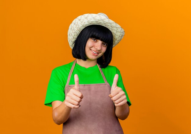 Sorridente giovane giardiniere femminile in uniforme che indossa il cappello da giardinaggio pollice in alto con due mani