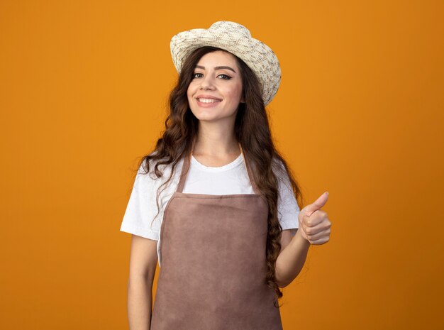 Sorridente giovane giardiniere femminile in uniforme che indossa il cappello da giardinaggio pollice in alto isolato sulla parete arancione con lo spazio della copia