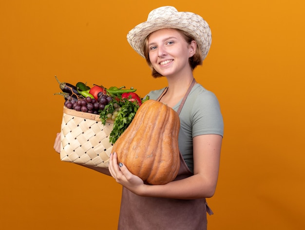 Sorridente giovane giardiniere femminile slavo indossando il cappello di giardinaggio tenendo il cesto di verdure e zucca sull'arancia