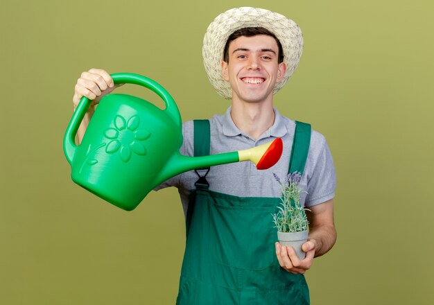Sorridente giovane giardiniere maschio che indossa il cappello da giardinaggio finge di innaffiare i fiori in vaso di fiori con annaffiatoio