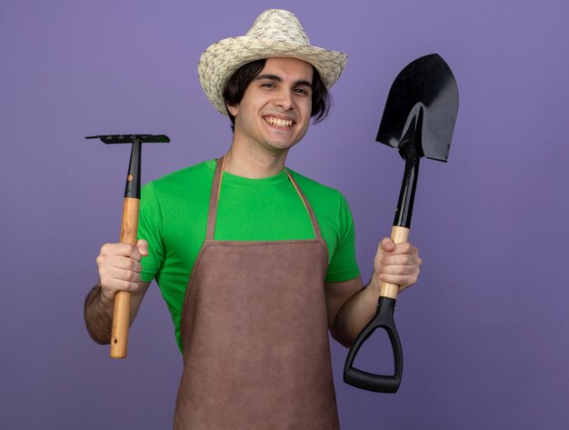 Sorridente giovane giardiniere maschio in uniforme che indossa cappello da giardinaggio tenendo il rastrello con vanga isolato su viola
