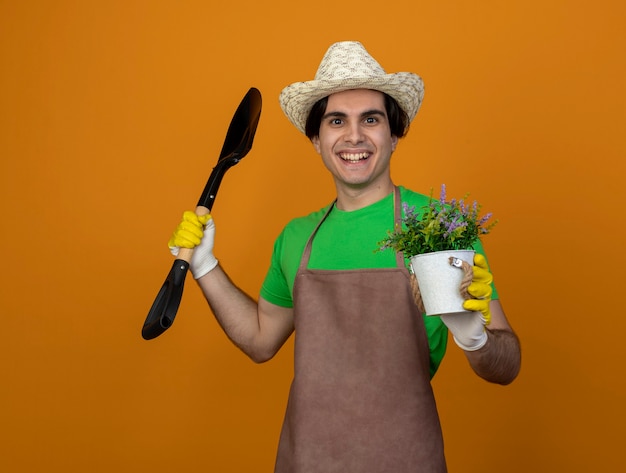 Sorridente giovane giardiniere maschio in uniforme che indossa il cappello da giardinaggio con guanti che tengono la vanga con il fiore in vaso di fiori isolato sulla parete arancione