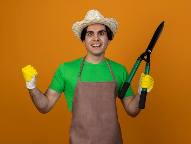 Sorridente giovane giardiniere maschio in uniforme che indossa il cappello di giardinaggio con i guanti che tengono i clippers mostrando sì gesto isolato sull'arancio