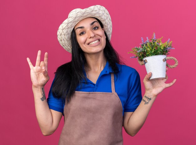 Sorridente giovane giardiniere ragazza che indossa l'uniforme e cappello tenendo il vaso di fiori guardando la parte anteriore facendo segno ok isolato sul muro rosa