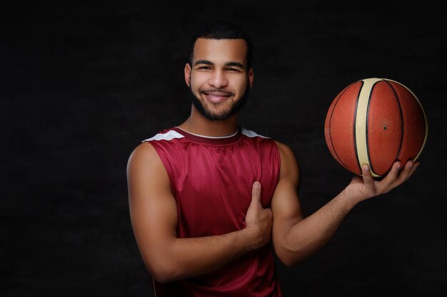 Sorridente giovane giocatore di basket afroamericano in abbigliamento sportivo su sfondo scuro.