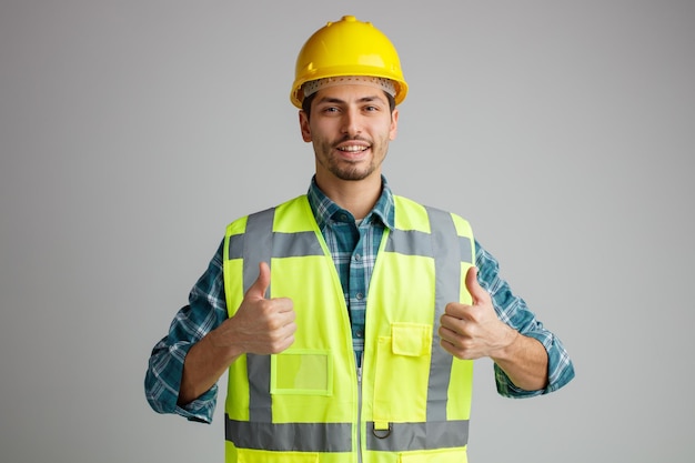 Sorridente giovane ingegnere maschio che indossa casco di sicurezza e uniforme guardando la fotocamera che mostra i pollici in su isolato su sfondo bianco