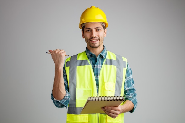Sorridente giovane ingegnere maschio che indossa un casco di sicurezza e uniforme che tiene blocco note e matita guardando la fotocamera che punta al lato isolato su sfondo bianco