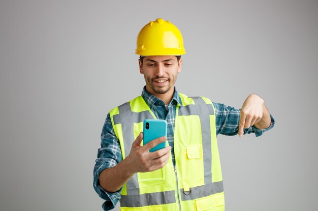Sorridente giovane ingegnere maschio che indossa un casco di sicurezza e uniforme che tiene e guarda il telefono cellulare rivolto verso il basso isolato su sfondo bianco