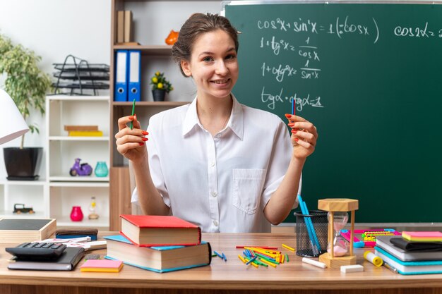 Sorridente giovane insegnante di matematica femminile seduto alla scrivania con forniture scolastiche tenendo bastoncini di conteggio guardando davanti in classe