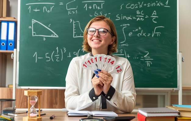 Sorridente giovane insegnante di sesso femminile con gli occhiali si siede a tavola con forniture scolastiche tenendo il numero di fan in classe