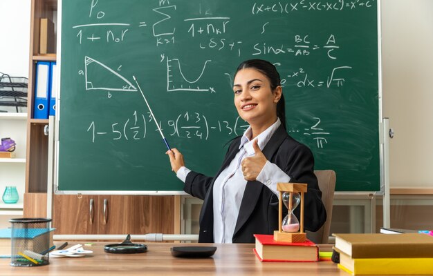 Sorridente giovane insegnante di sesso femminile si siede al tavolo con forniture scolastiche punti alla lavagna con il bastone puntatore che mostra il pollice in su in classe