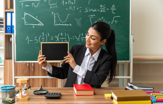 Sorridente giovane insegnante femminile si siede al tavolo con gli strumenti della scuola che tengono e guardano la mini lavagna in classe