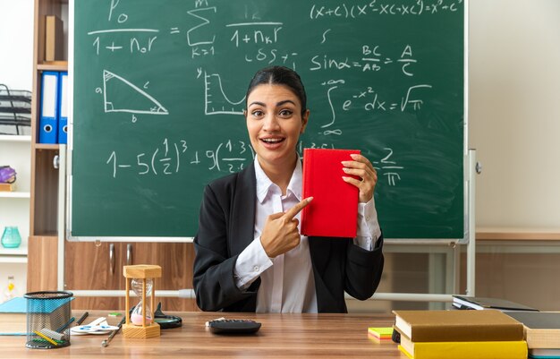 Sorridente giovane insegnante femminile si siede al tavolo con materiale scolastico in mano e indica i libri in classe