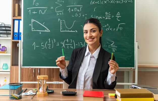 Sorridente giovane insegnante femminile si siede al tavolo con materiale scolastico tenendo la matita in classe