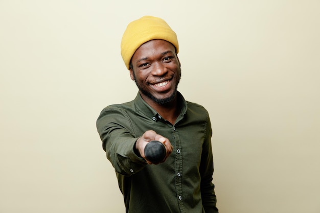 Sorridente giovane maschio afroamericano con cappello che indossa una camicia verde che porge il microfono alla telecamera isolato su sfondo bianco