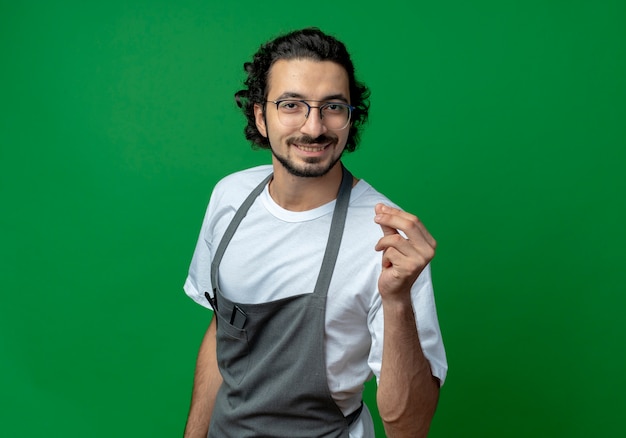 Sorridente giovane maschio caucasico barbiere con gli occhiali e fascia per capelli ondulati in uniforme che fa gesto di denaro isolato su priorità bassa verde con lo spazio della copia