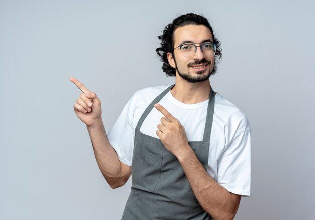 Sorridente giovane maschio caucasico barbiere con gli occhiali e fascia per capelli ondulati in uniforme che punta a lato isolato su sfondo bianco