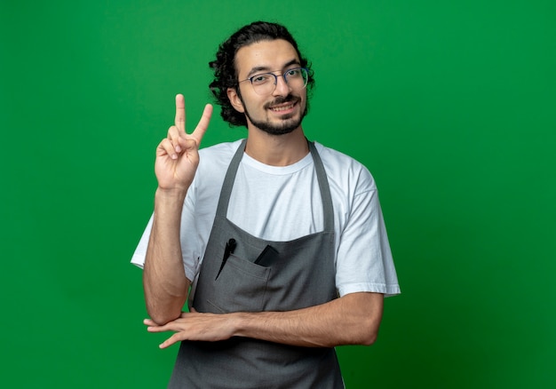 Sorridente giovane maschio caucasico barbiere con gli occhiali e fascia per capelli ondulati in uniforme facendo segno di pace e mettendo la mano sotto il gomito isolato su sfondo verde con spazio di copia