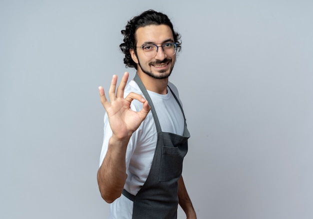 Sorridente giovane maschio caucasico barbiere con gli occhiali e fascia per capelli ondulati in uniforme in piedi in vista di profilo facendo segno ok