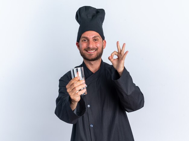 Sorridente giovane maschio caucasico cuoco in uniforme da chef e berretto che allunga un bicchiere d'acqua verso la telecamera guardando la telecamera facendo segno ok isolato sul muro bianco con spazio di copia