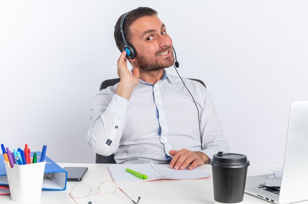 Sorridente giovane maschio operatore di call center che indossa la cuffia seduto al tavolo con strumenti per ufficio isolato sul muro bianco