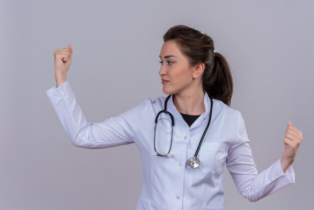 Sorridente giovane medico che indossa abito medico indossando stetoscopio pugno alzato sul muro bianco