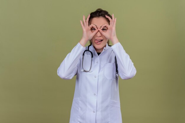 Sorridente giovane medico che indossa abito medico indossando uno stetoscopio che mostra il gesto di sguardo sulla parete verde