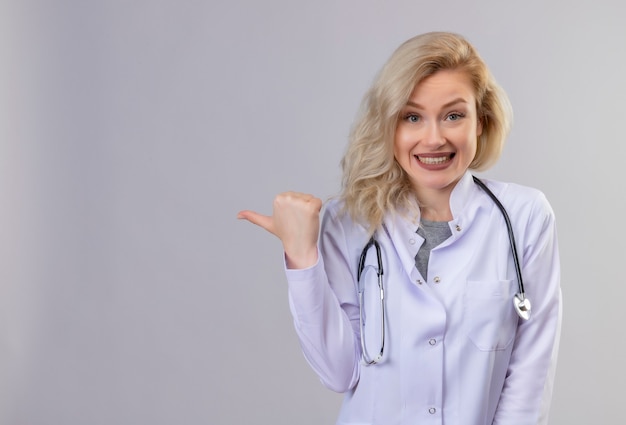 Sorridente giovane medico che indossa lo stetoscopio in abito medico punta a lato sul muro bianco