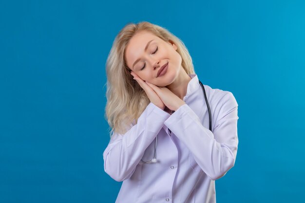 Sorridente giovane medico che indossa uno stetoscopio in abito medico che mostra il gesto del sonno sulla parete blu