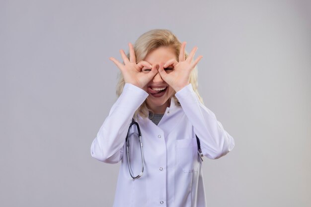 Sorridente giovane medico che indossa uno stetoscopio in abito medico che mostra il gesto di sguardo sulla parete bianca