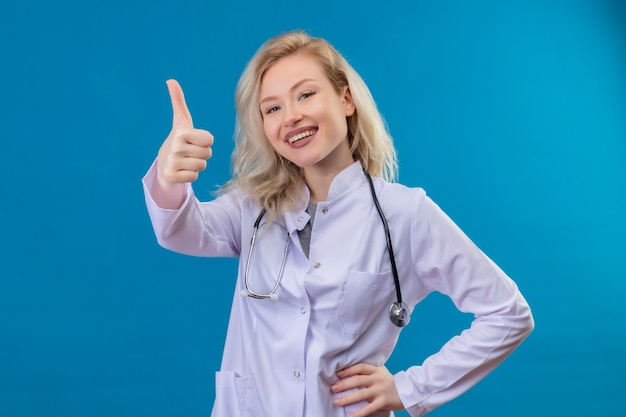 Sorridente giovane medico che indossa uno stetoscopio in abito medico il suo pollice in alto sulla parete blu