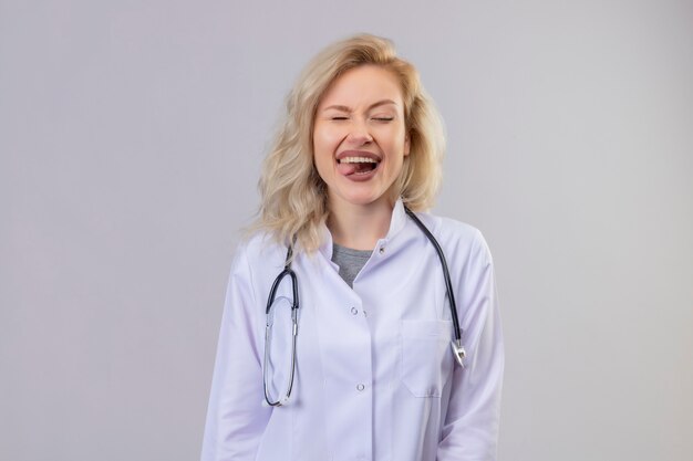 Sorridente giovane medico che indossa uno stetoscopio in abito medico morde la lingua sul muro bianco