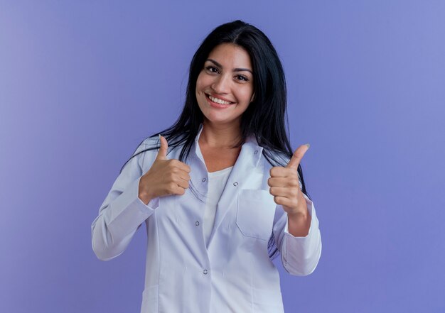 Sorridente giovane medico femminile che indossa abito medico cercando di mostrare i pollici