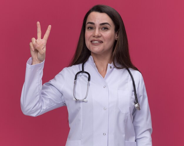Sorridente giovane medico femminile che indossa abito medico con gesti di stetoscopio mano segno di vittoria sul rosa