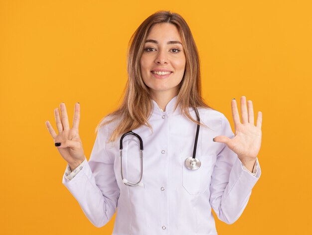 Sorridente giovane medico femminile che indossa abito medico con lo stetoscopio che mostra i numeri differenti isolati sulla parete gialla