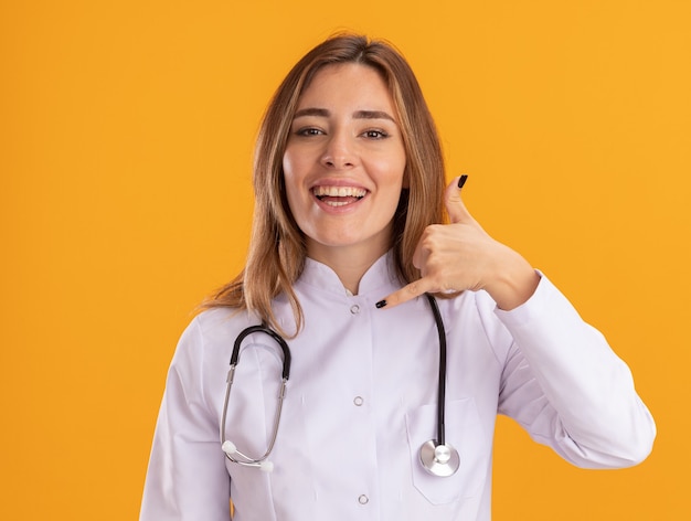 Sorridente giovane medico femminile che indossa abito medico con lo stetoscopio che mostra il gesto di chiamata telefonica isolato sulla parete gialla