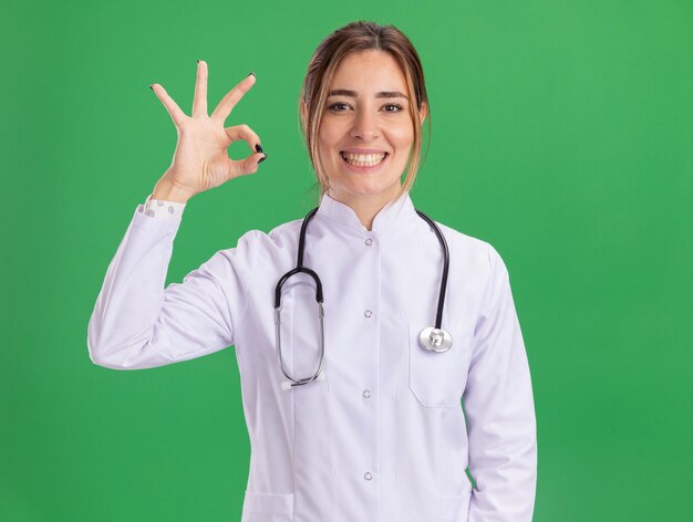 Sorridente giovane medico femminile che indossa abito medico con lo stetoscopio che mostra il gesto giusto isolato sulla parete verde