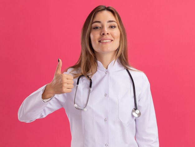 Sorridente giovane medico femminile che indossa abito medico con lo stetoscopio che mostra il pollice in alto isolato sulla parete rosa