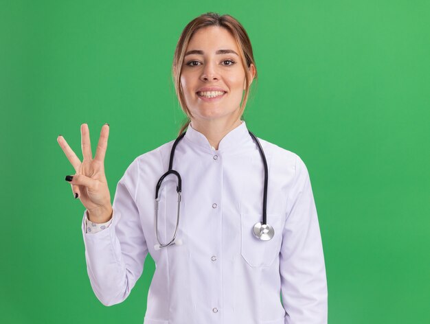 Sorridente giovane medico femminile che indossa abito medico con uno stetoscopio che mostra tre isolati sulla parete verde