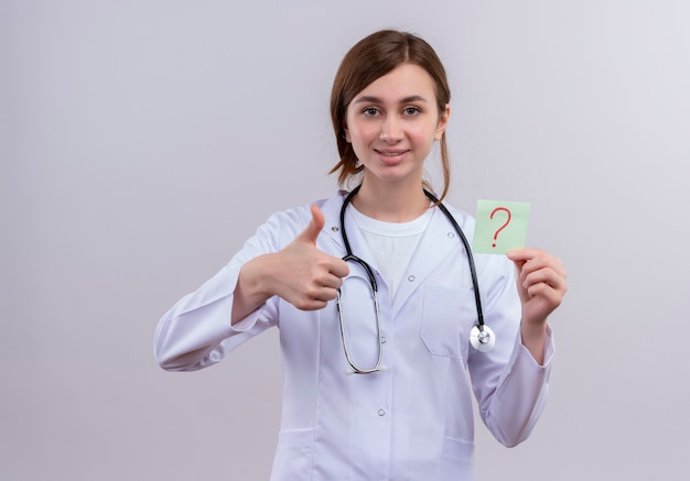 Sorridente giovane medico femminile che indossa abito medico e stetoscopio che mostra pollice in alto e che tiene nota di carta con il punto interrogativo sulla parete bianca isolata