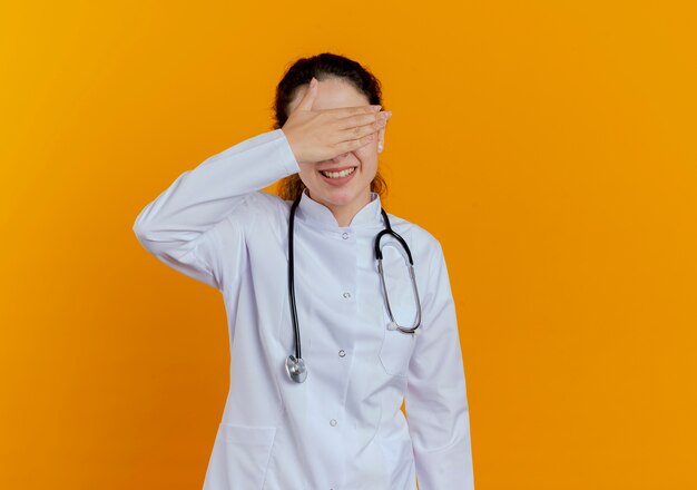 Sorridente giovane medico femminile che indossa abito medico e stetoscopio con gli occhiali coperti con gli occhi della mano isolati