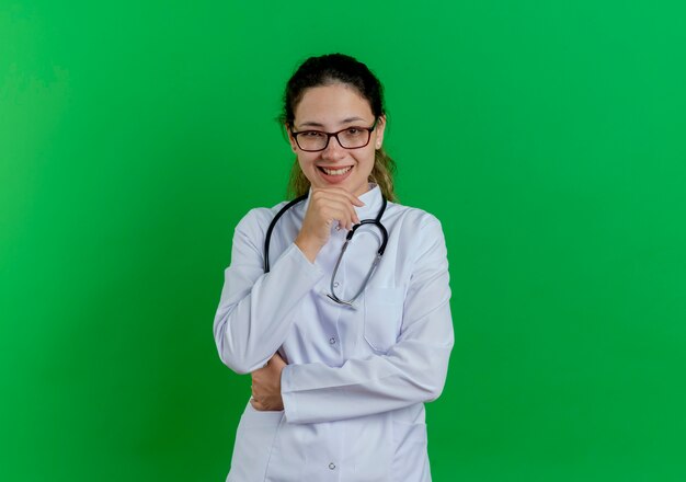 Sorridente giovane medico femminile che indossa abito medico e stetoscopio e bicchieri toccando il mento isolato sulla parete verde con lo spazio della copia