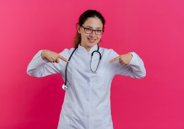 Sorridente giovane medico femminile che indossa abito medico e stetoscopio e occhiali che punta a se stessa isolato sulla parete rosa