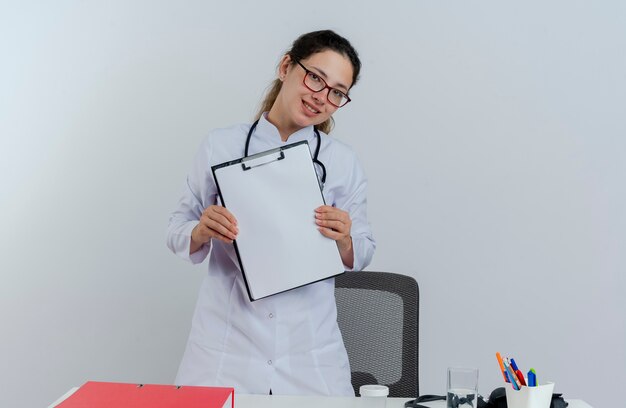 Sorridente giovane medico femminile che indossa abito medico e stetoscopio e occhiali in piedi dietro la scrivania con strumenti medici che sembrano mostrare appunti isolati