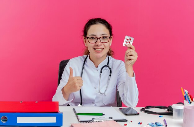 Sorridente giovane medico femminile che indossa abito medico e stetoscopio e occhiali seduto alla scrivania